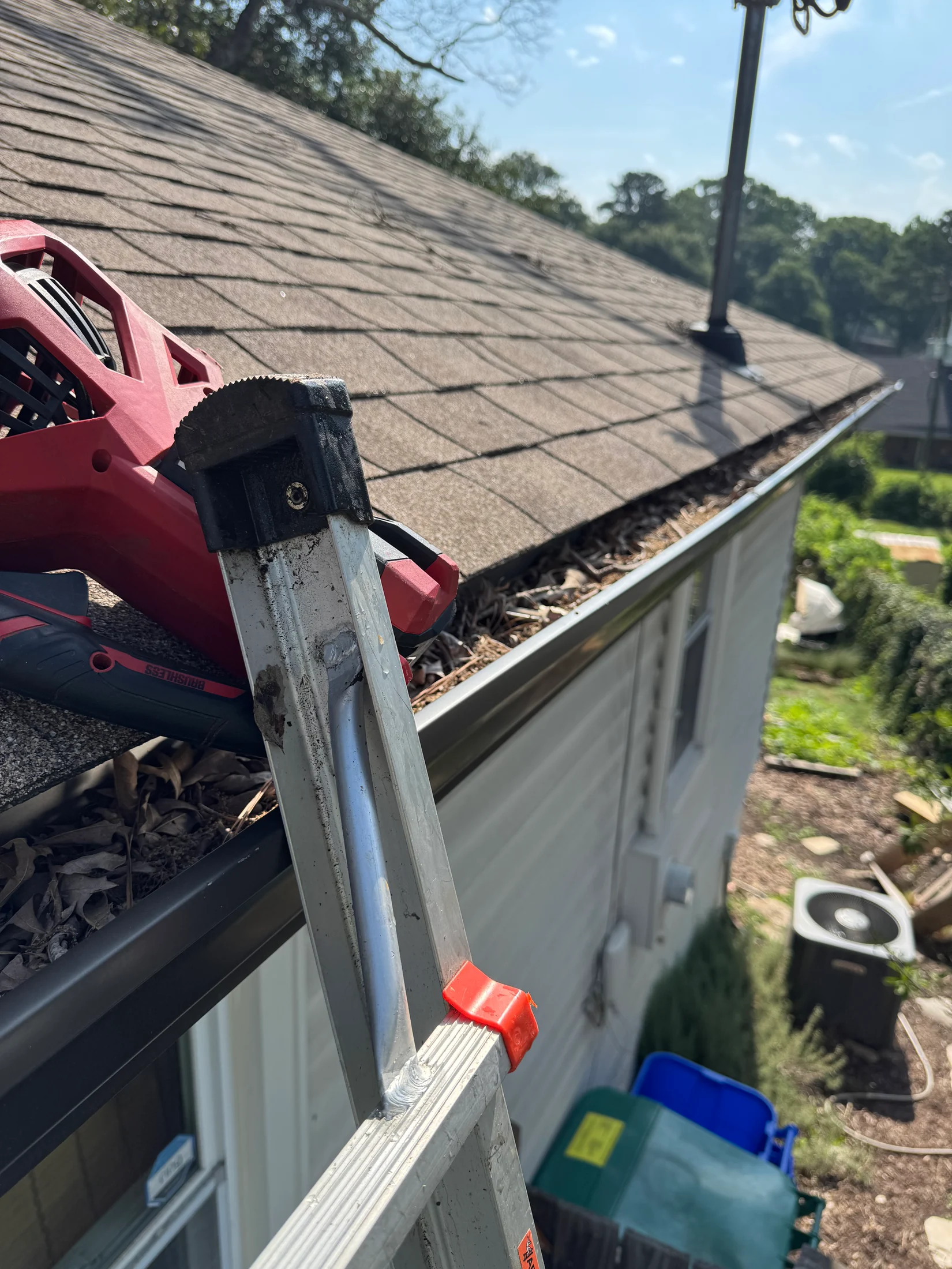 An extension ladder and blower beside a gutter containing leaves at the roofline.