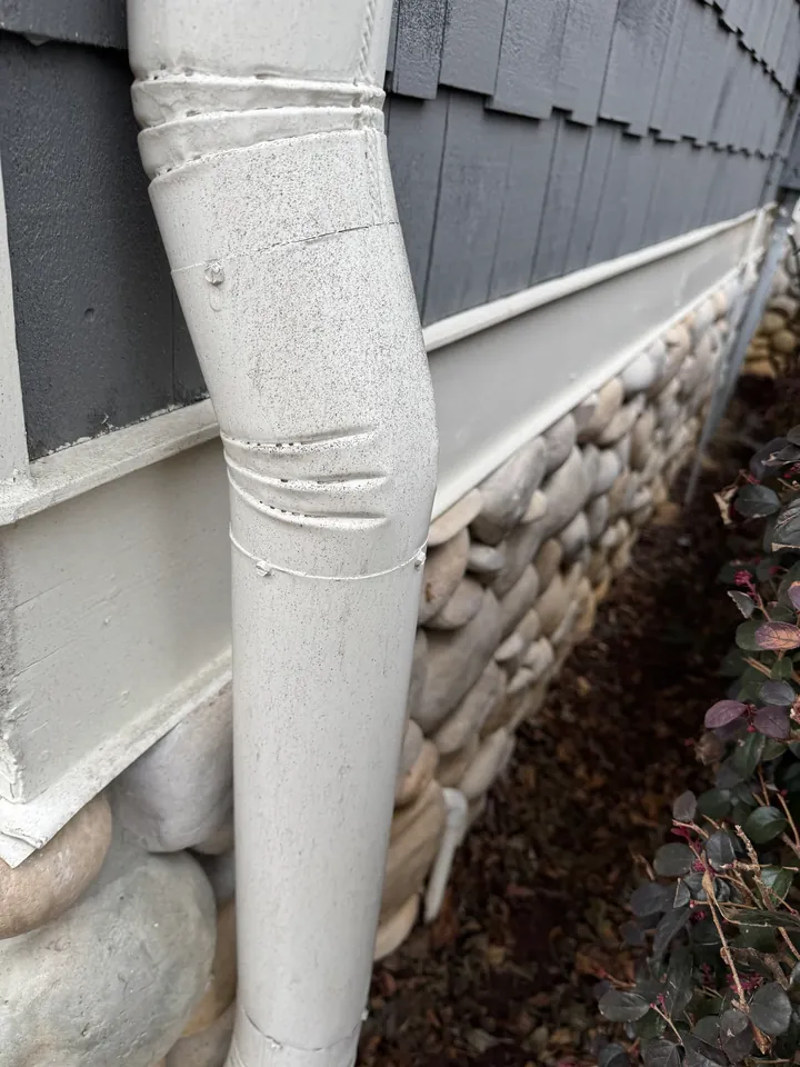 White downspout beside gray siding, white trim and a stone-clad foundation.