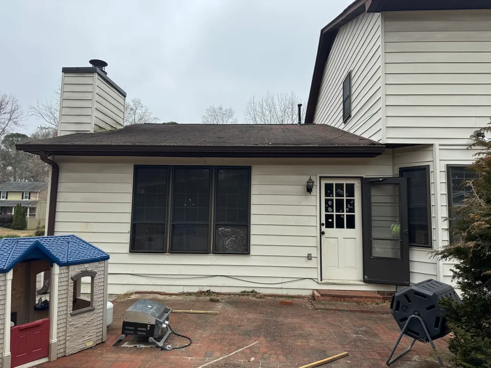 Rear house elevation with cream siding, dark shutters, a brick patio, a hose and cleaning equipment.